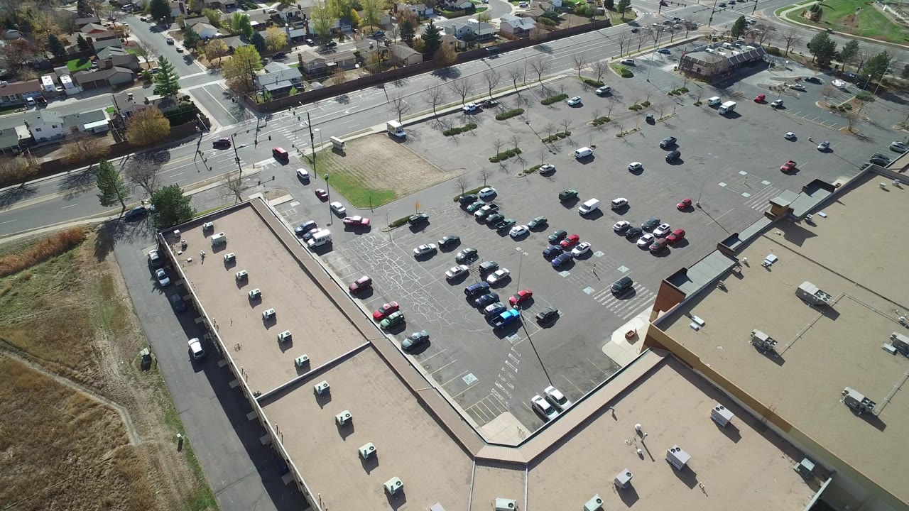 Opening aerial frame over a flat-roof commercial building and large parking lot in Hidden Lake, Colorado, with scattered cars, surrounding roads, residential blocks, and no visible people.