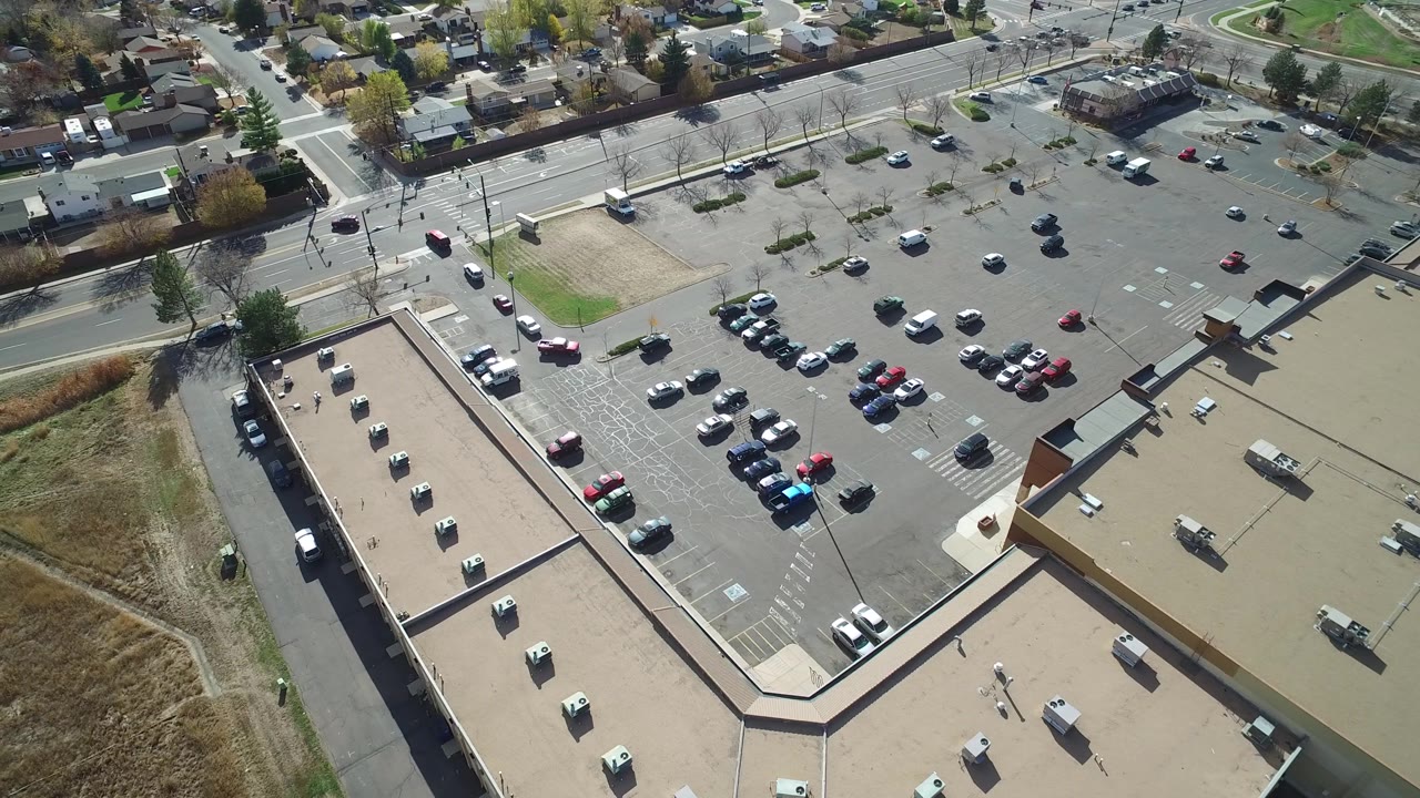 Aerial frame in Hidden Lake, Colorado showing the commercial roof at lower left, wide parking lanes with scattered vehicles, road traffic at the top, and no visible people.