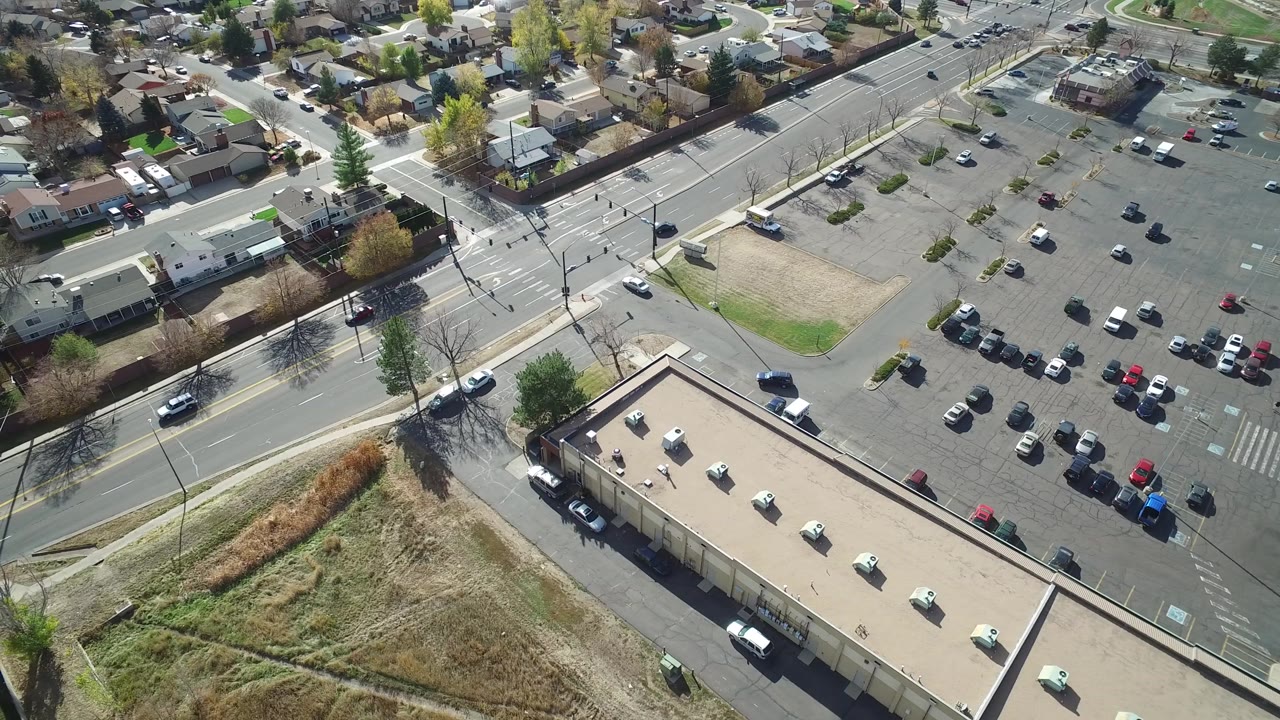 Drone view in Hidden Lake, Colorado with the road junction near center, a large parking lot to the right, houses beyond the streets, dry grass below, and no visible people.