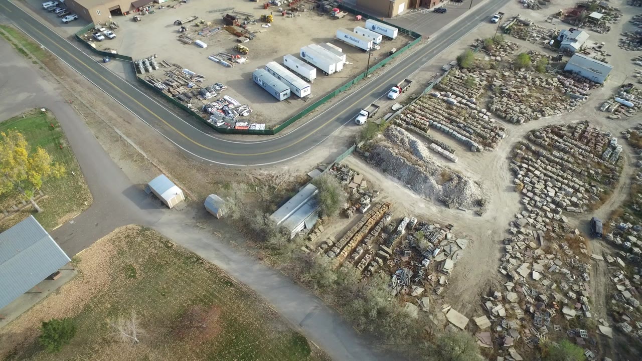 Aerial view of a curved road beside storage yards, stacked materials, sheds, and dry grass.