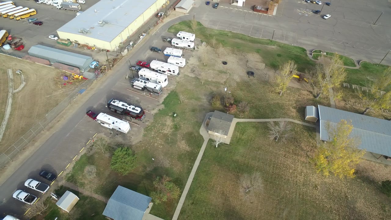Aerial frame over Hidden Lake, Colorado with white trailers clustered near parked vehicles, a pale-roofed building, brown-green grass, roads, and tiny possible people too small to identify.