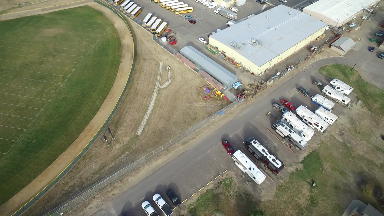 High aerial view of a baseball field beside a service road and parked vehicles.