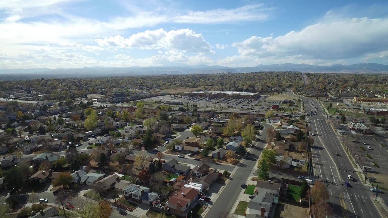 Aerial frame over Hidden Lake, Colorado showing residential roofs, tree-lined blocks, a multi-lane road, distant mountains, scattered clouds, and no visible people.