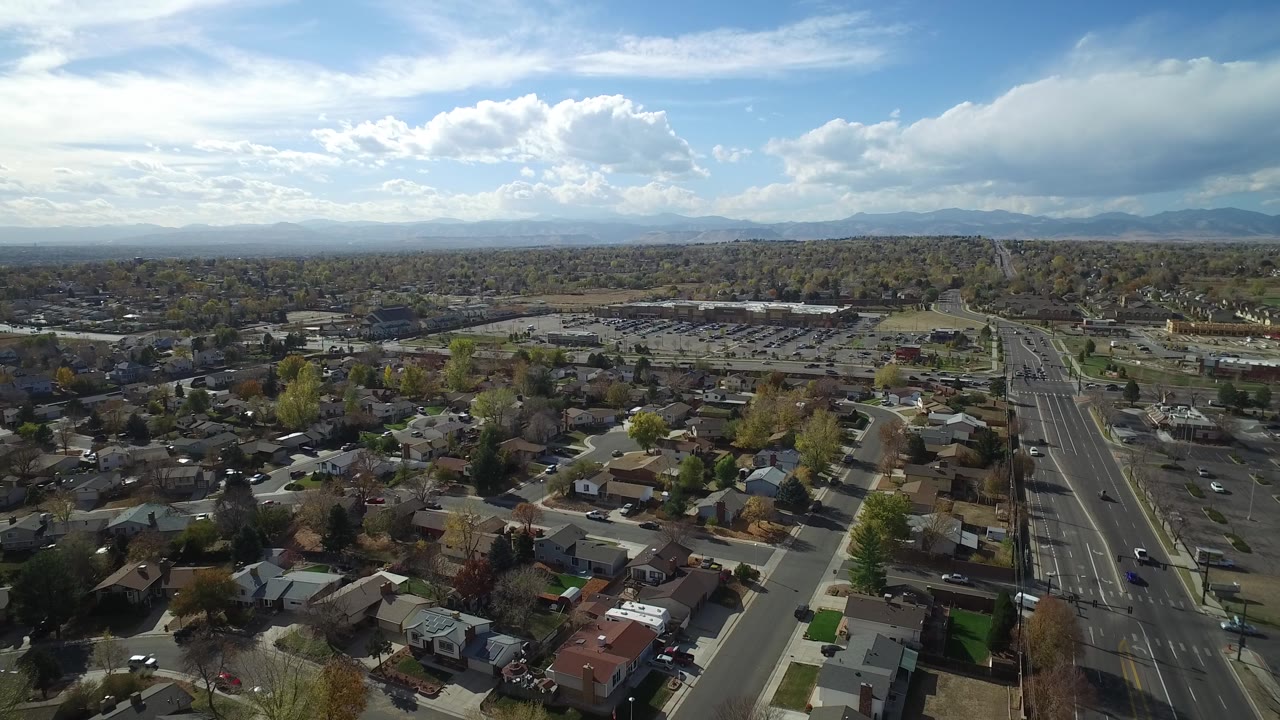 Suburban neighborhood aerial with a broad road and mountains beneath large clouds.