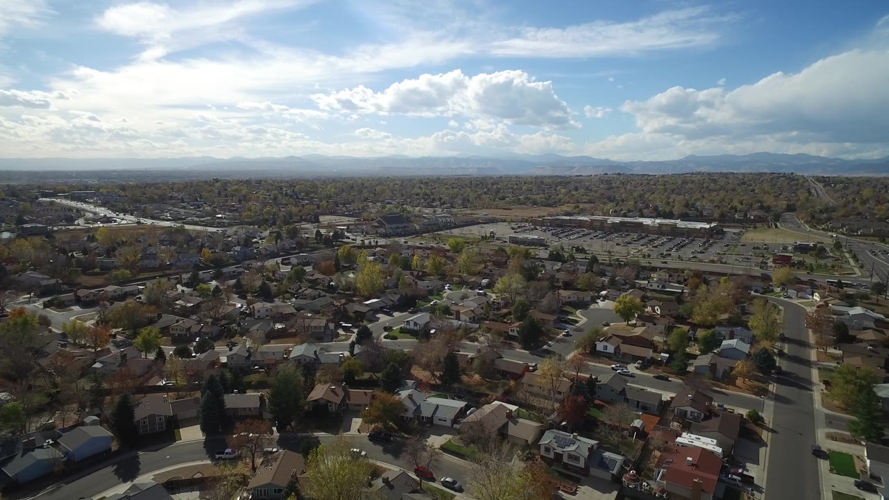 Wide aerial frame over Hidden Lake, Colorado showing residential rooftops, a straight neighborhood road, autumn trees, low hills, distant mountains, bright sky, and no visible people.