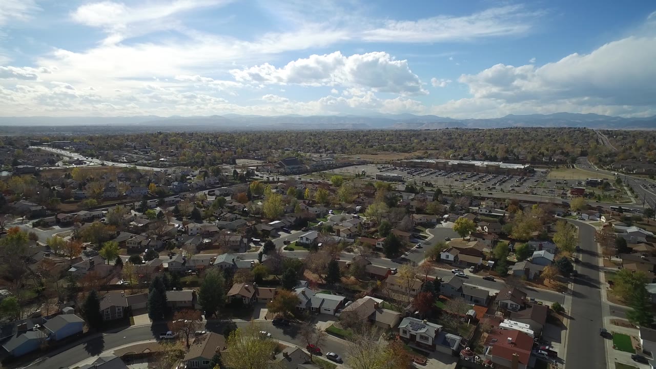 Aerial view over Hidden Lake, Colorado with houses and tree-lined streets in the lower frame, a broad developed area beyond, mountains on the horizon, and no visible people.