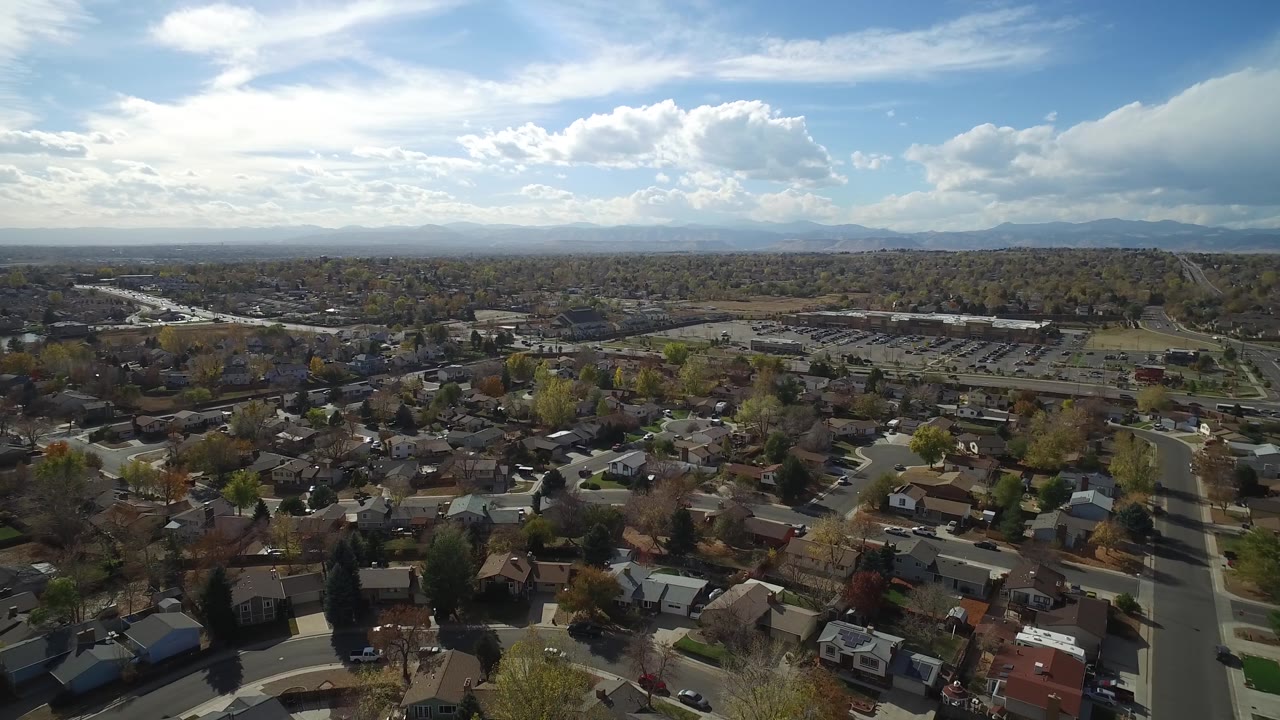 Drone-height view of rooftops, roads, parking area, hills, and mountains.