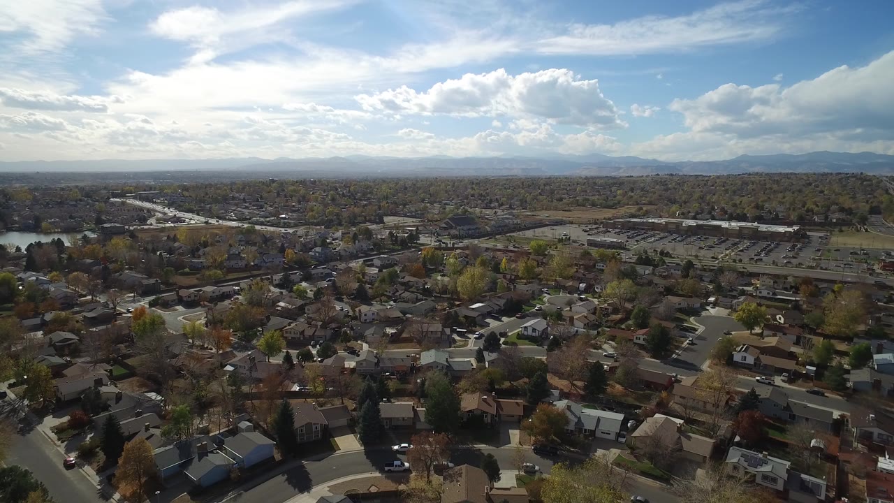 Aerial view of homes, trees, lake water, and distant mountains under a cloudy blue sky.