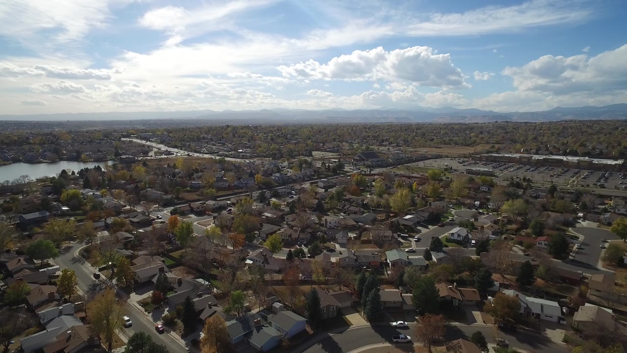 Opening aerial frame over Hidden Lake, Colorado with suburban rooftops in the foreground, a small lake to the left, distant mountains, bright cloud cover, and no visible people.