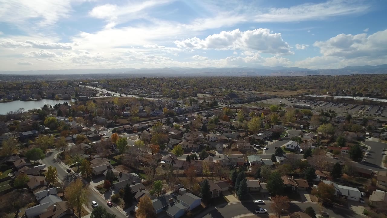 Drone-height view across rooftops, streets, lake edge, trees, and mountains beneath a blue sky.