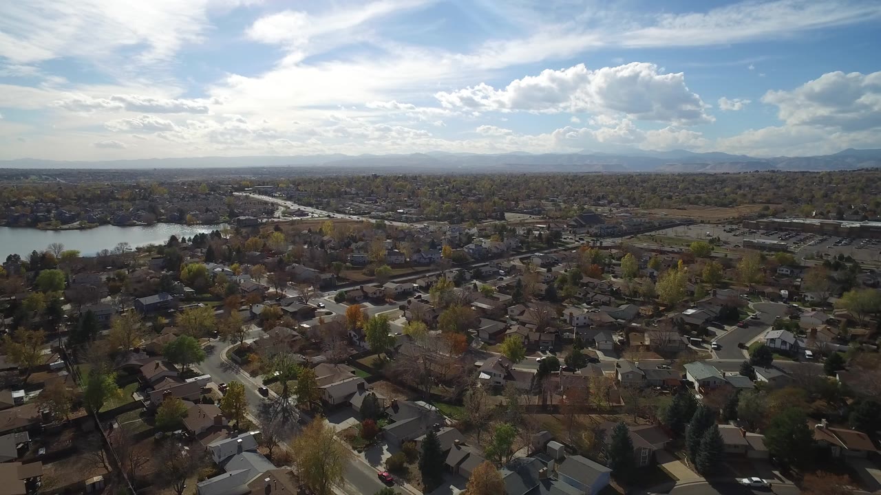 Aerial view of homes, trees, lake, roads, and distant mountains under clouds.