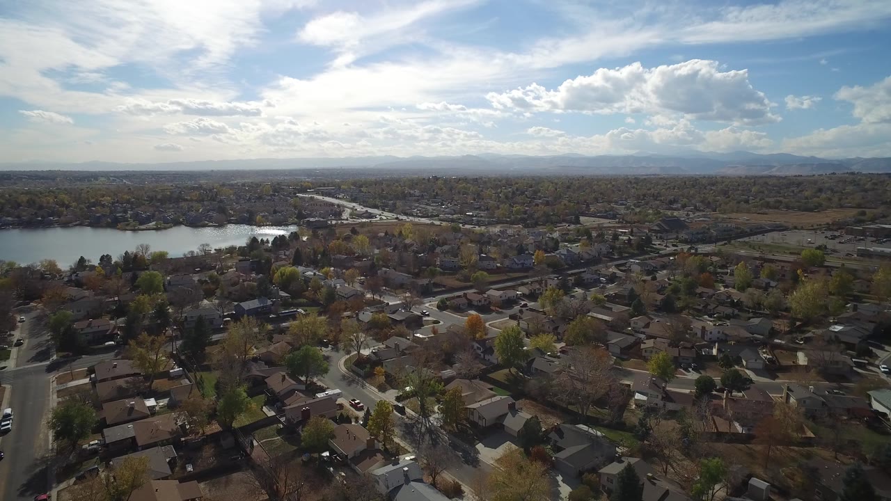 Aerial frame over Hidden Lake, Colorado with rooftops and intersecting streets below, a lake running across the midground, hazy mountains on the horizon, high clouds, and no visible people.