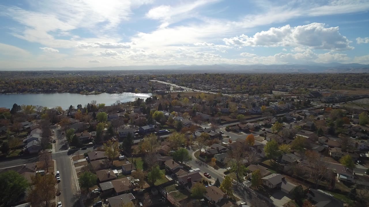 Aerial view of homes, streets, a lake, and distant mountains under a bright sky.
