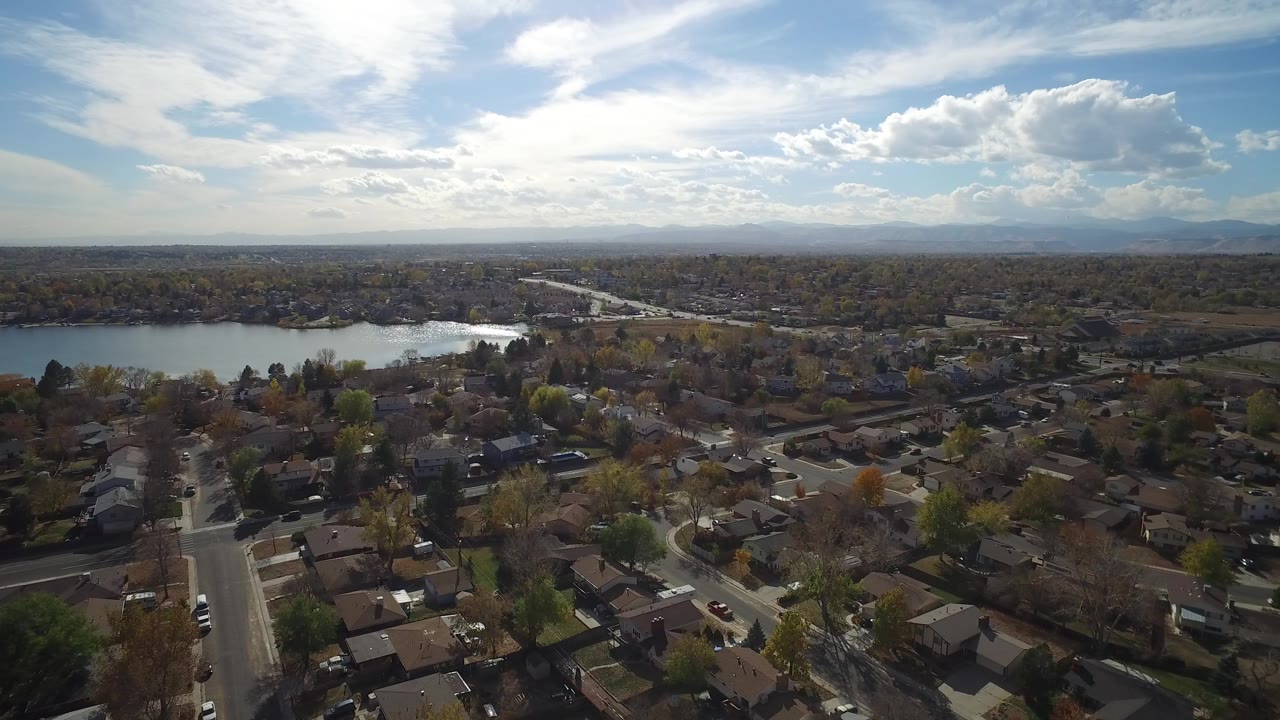 Early aerial frame in Hidden Lake, Colorado, looking over a straight residential street, clustered homes, lakeside trees, reflective water, distant hills, and no visible people.