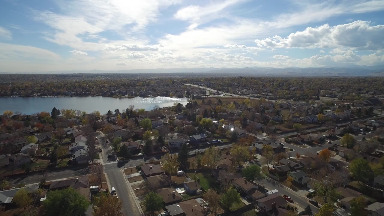 Aerial view of suburban homes, streets, trees, lake, and bright clouded sky.