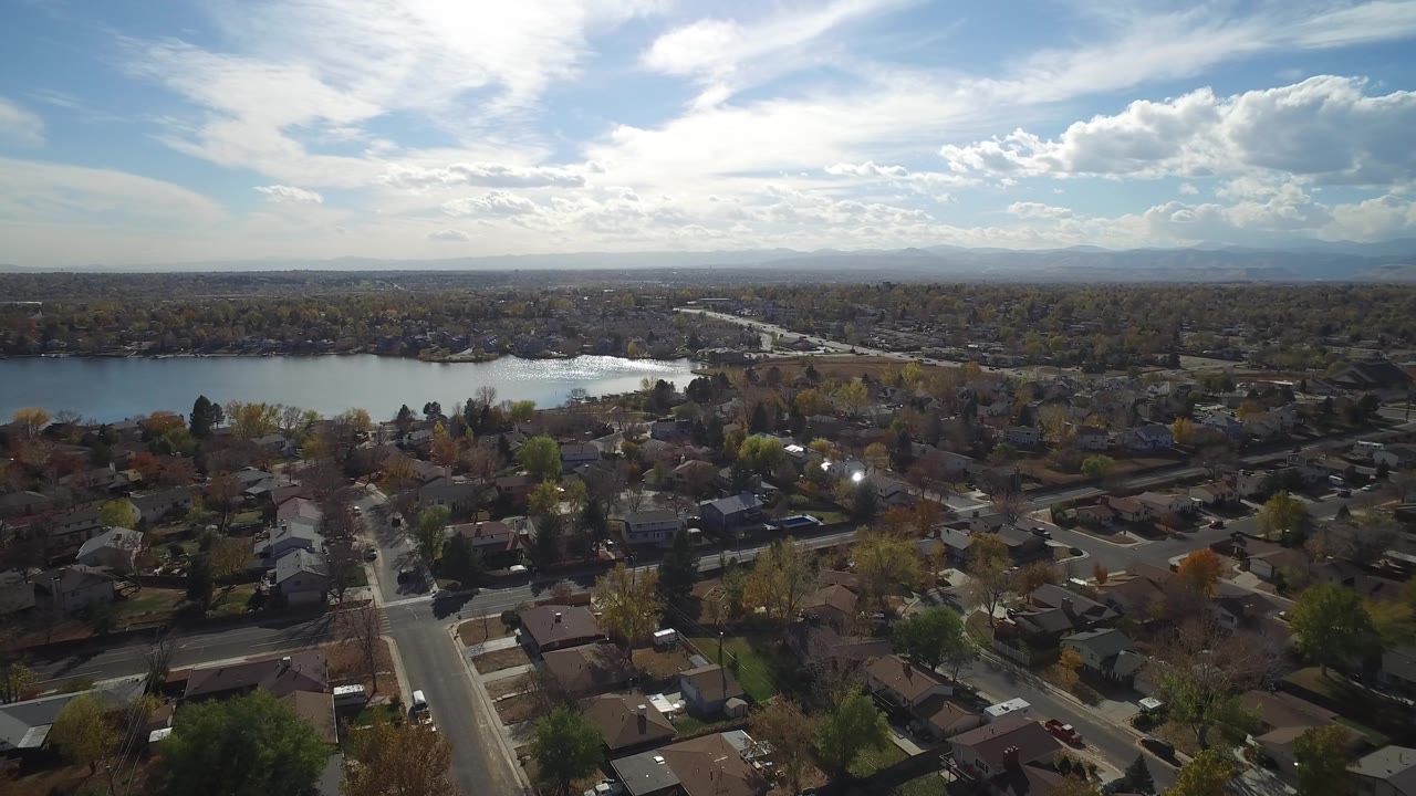 Second aerial frame in Hidden Lake, Colorado, with rooftops and tree-lined blocks below, the lake across the midground, strong daylight glare, and no visible people.