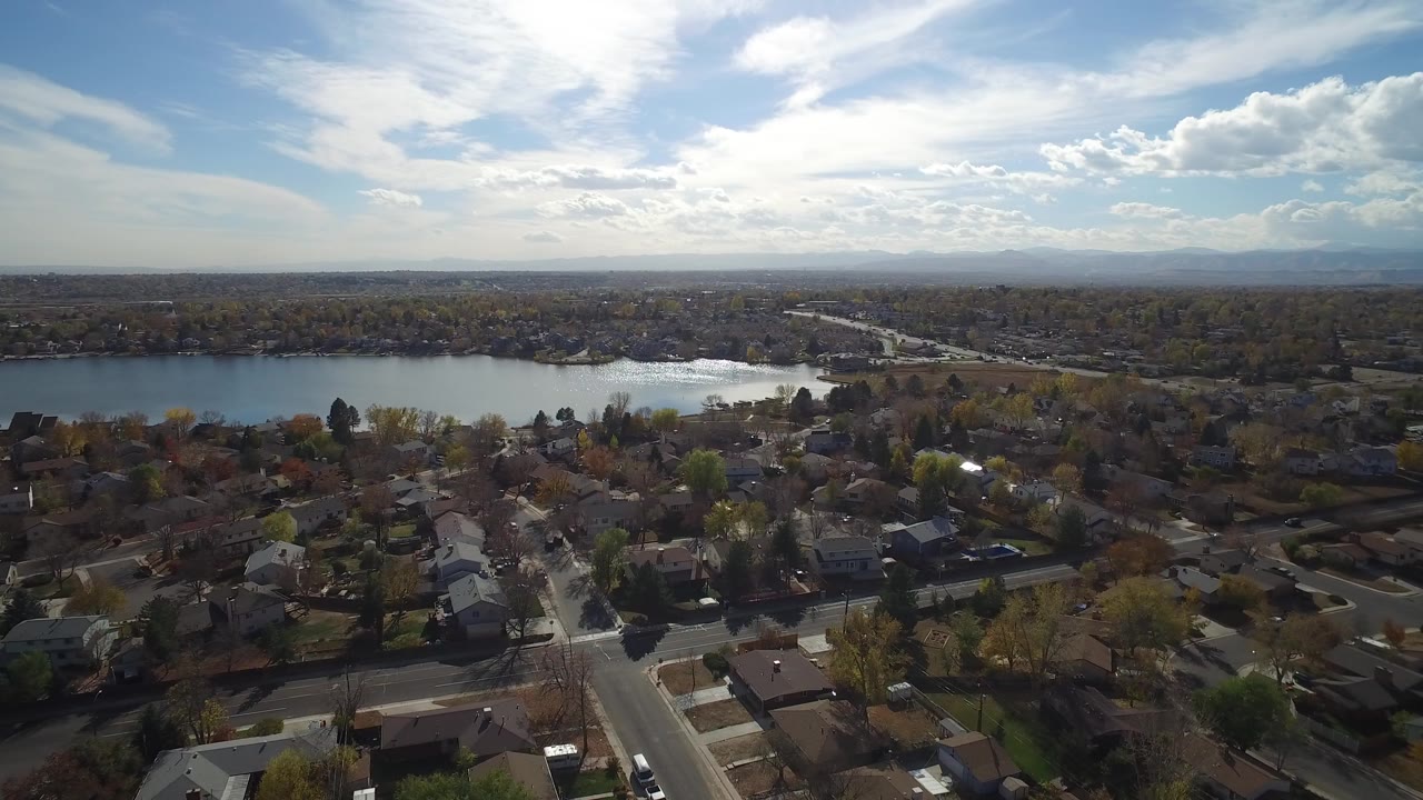 Aerial view of houses, streets, trees, lake, and bright sky.