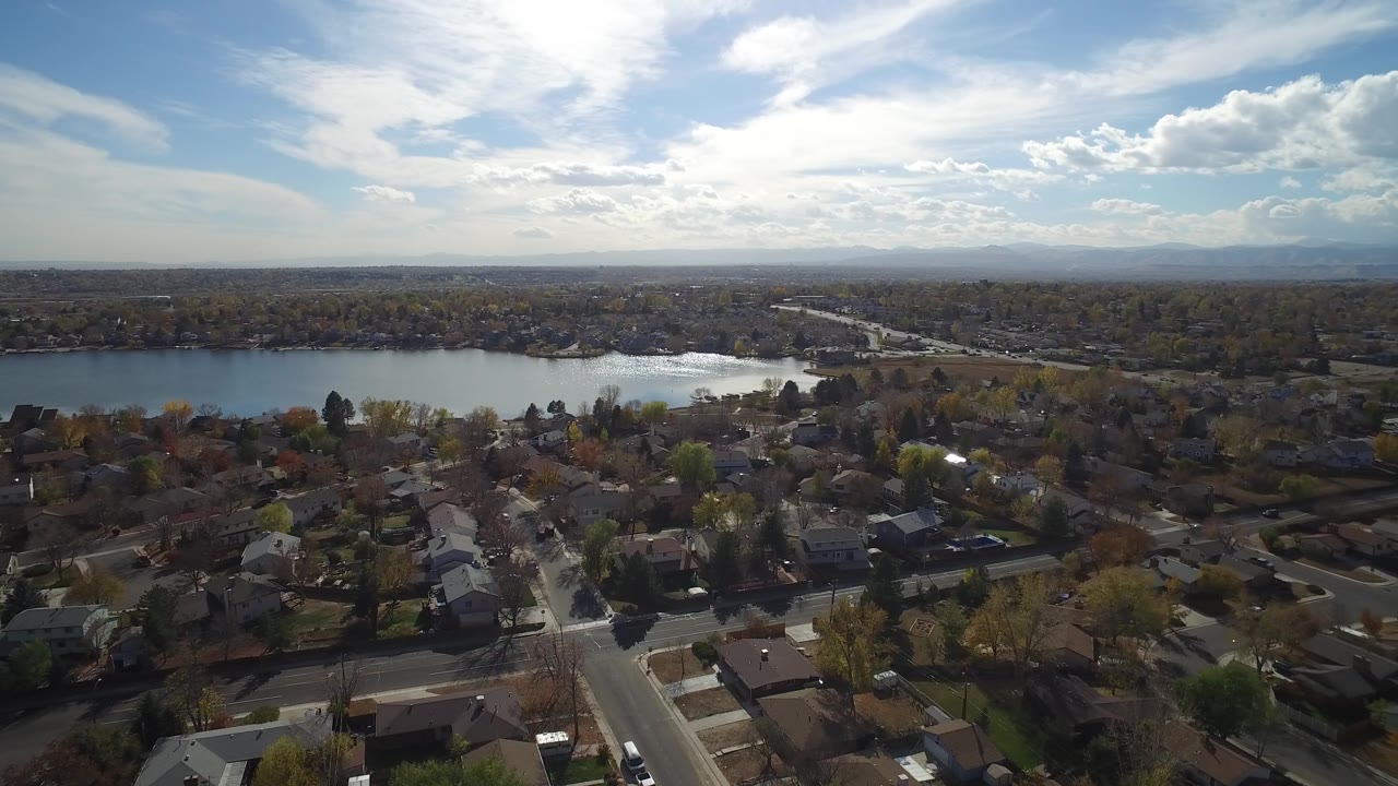 Aerial frame over Hidden Lake, Colorado showing the lake behind a spread of roofs, yards, streets, leafed and bare trees, a bright cloud bank, and no visible people.