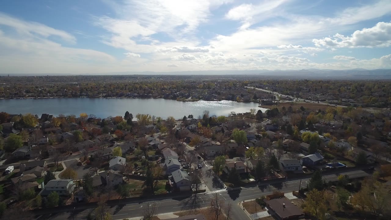 Aerial view of homes, streets, lake, and distant horizon under bright clouds.