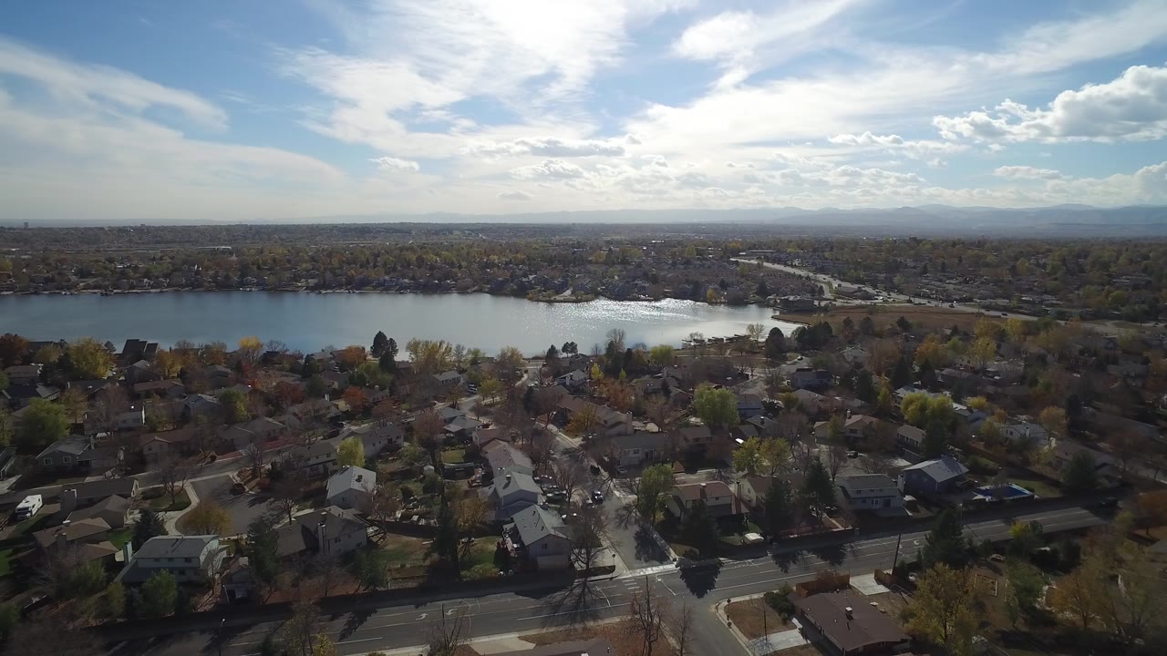 Second aerial frame of Hidden Lake, Colorado showing tree-lined residential blocks below, the lake running horizontally through the scene, distant development on the horizon, and no visible people.