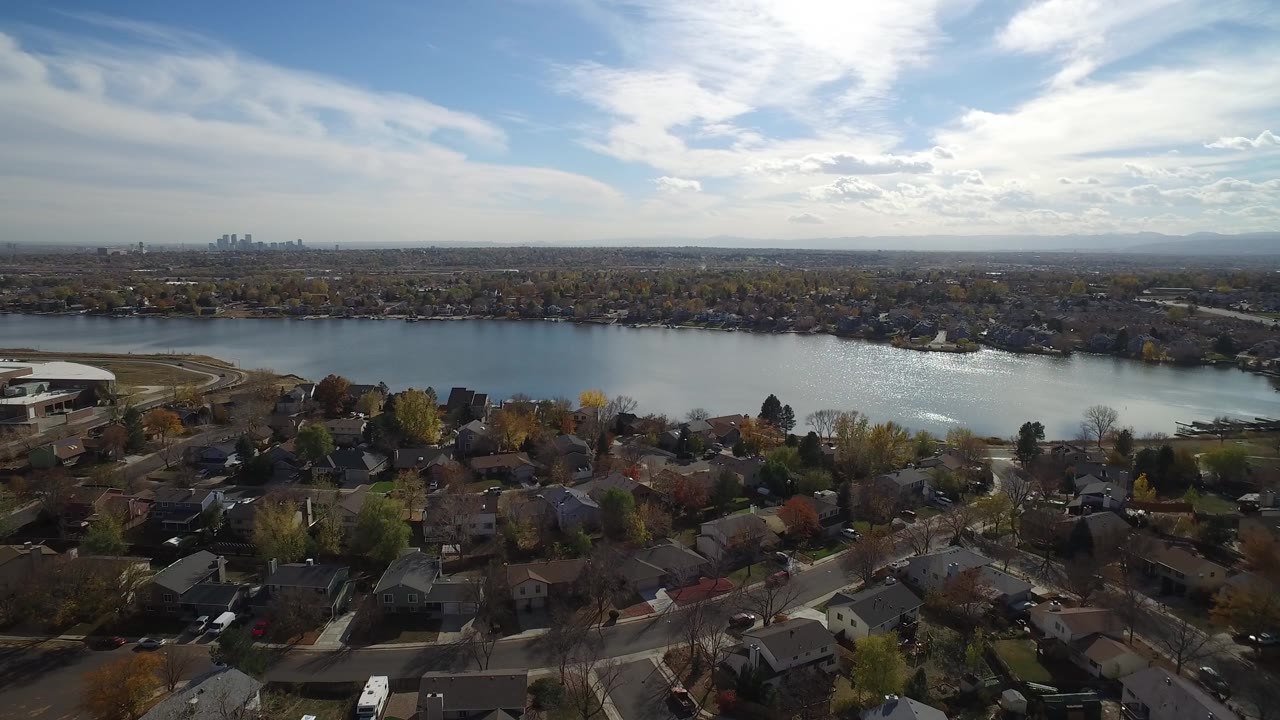 Aerial view of rooftops, trees, water, and a distant skyline under bright clouds.