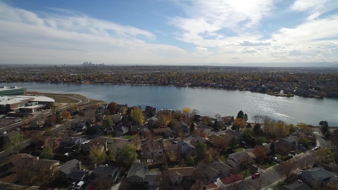 Wide aerial view over Hidden Lake, Colorado with residential rooftops in the foreground, a large light-roofed facility on the left shore, reflective lake water, distant towers, and no visible people.