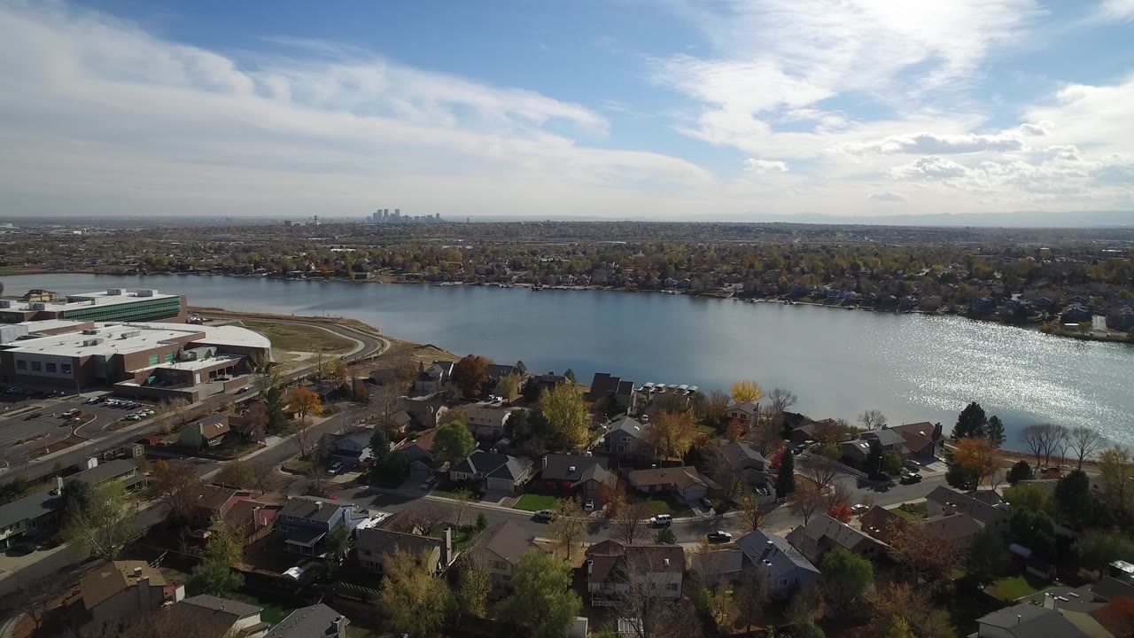 Hidden Lake, Colorado aerial frame with a lakeside neighborhood below, a large light-roofed building near the shore, sun reflection on the water, far skyline, and no visible people.