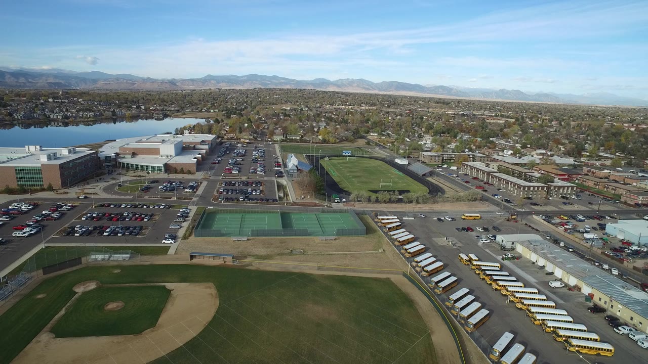 Opening aerial frame over Hidden Lake, Colorado with a baseball diamond in the lower left, tennis courts and school buses near the center, a lake to the left, distant mountains on the horizon, and no visible people.