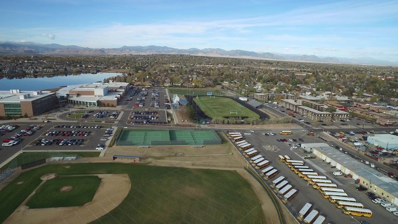 High drone view of baseball diamond, buses, roads, parking areas, lake, and suburban homes.