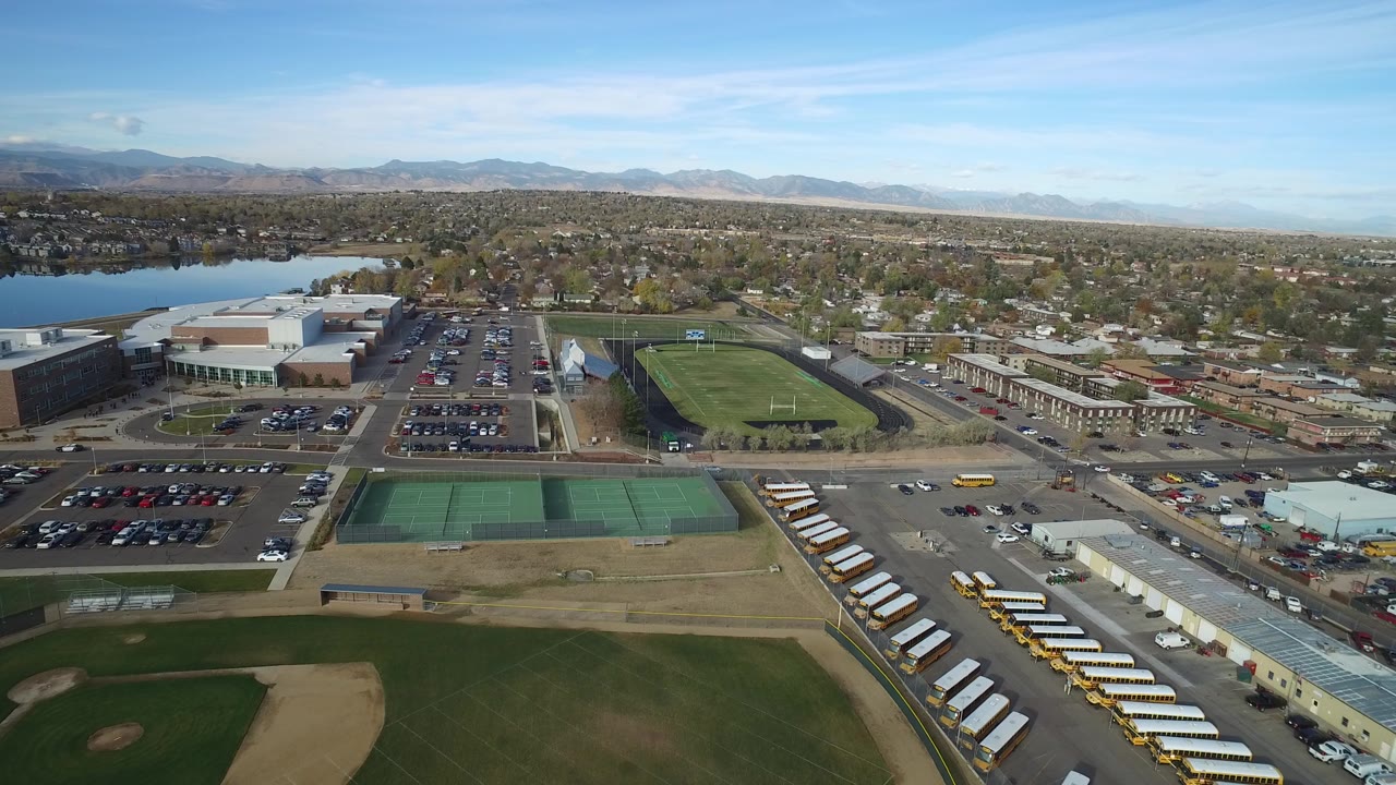 Aerial view over sports fields, parking lots, buses, a lake, and distant neighborhoods.