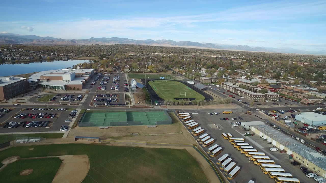 Hidden Lake, Colorado aerial still with green tennis courts and a baseball field low in frame, yellow school buses along the right side, parking lots near large buildings, a lake to the left, and no visible people.