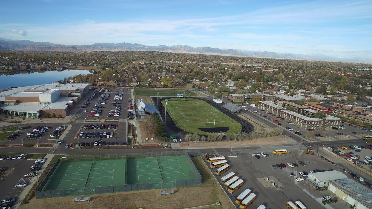 Opening aerial frame over Hidden Lake, Colorado showing green tennis courts at the bottom, school buses along a road, a black running track around a field, homes, a lake, distant mountains, and no visible people.