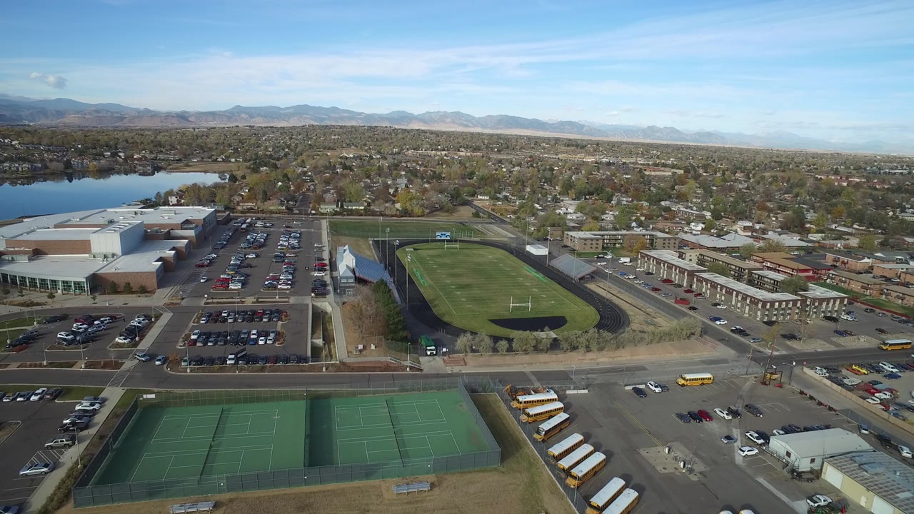 Aerial suburban view with tennis courts, school buses, a track field, roads, lake, homes, and mountains.
