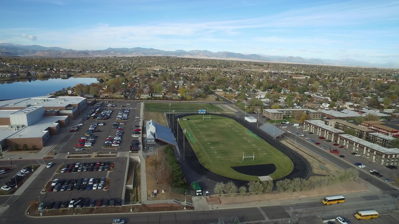 Aerial view of a school athletic field, parking lots, homes, lake, and mountains.