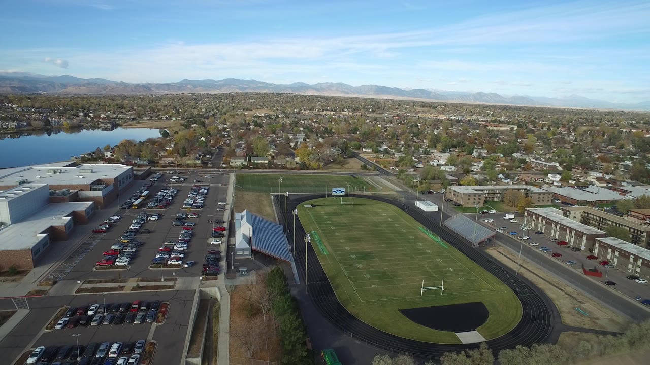 Aerial view of a school football field, track, parking lots, lake, homes, and mountains.