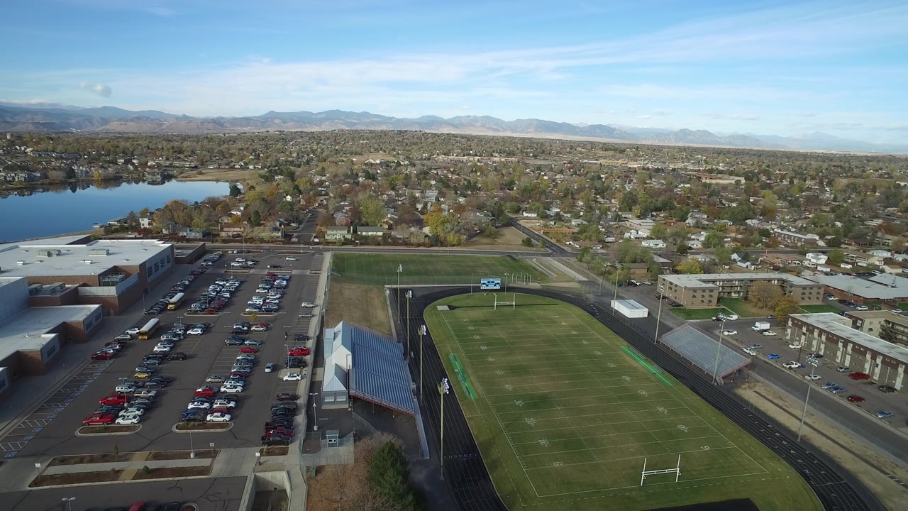 Aerial view of a school football field, parking lot, lake, homes, and distant mountains.