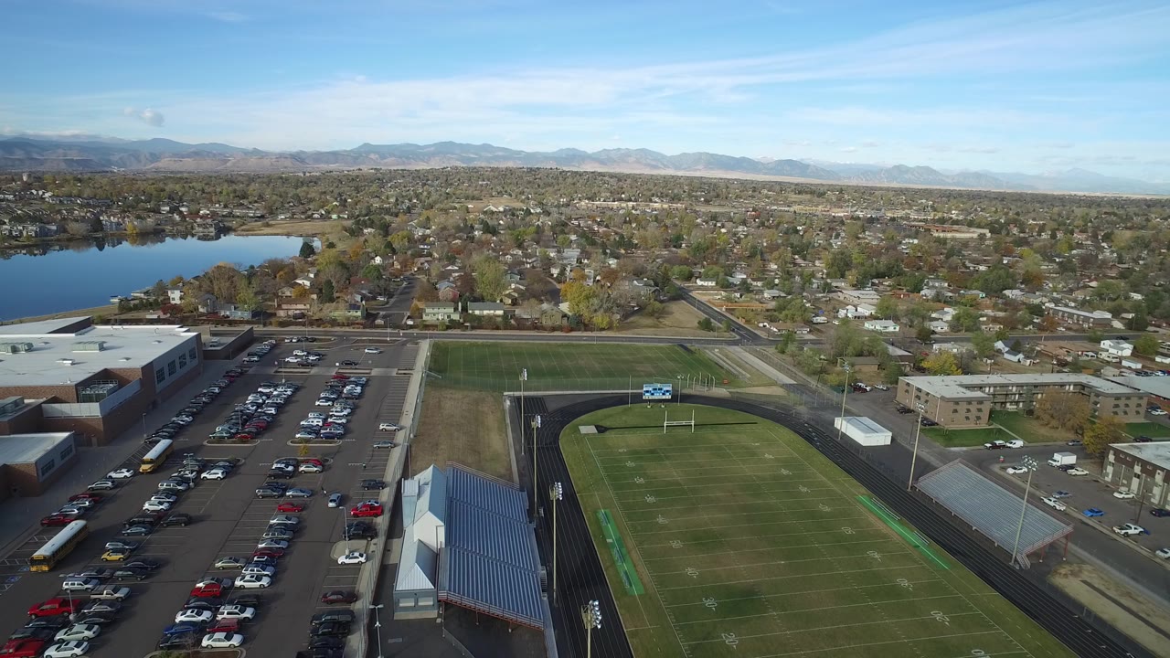Aerial view over a football field, parking lot, homes, lake, and distant mountains.