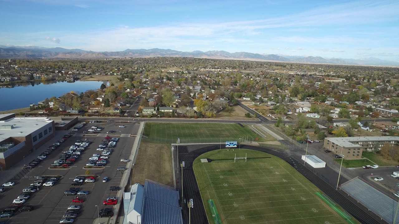Aerial view of a football field, parking lot, lake, neighborhood, and distant mountains.