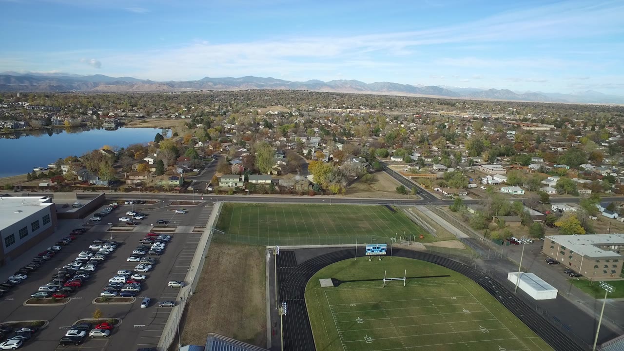 Aerial view of a football field, parking lots, homes, lake, and distant mountains.