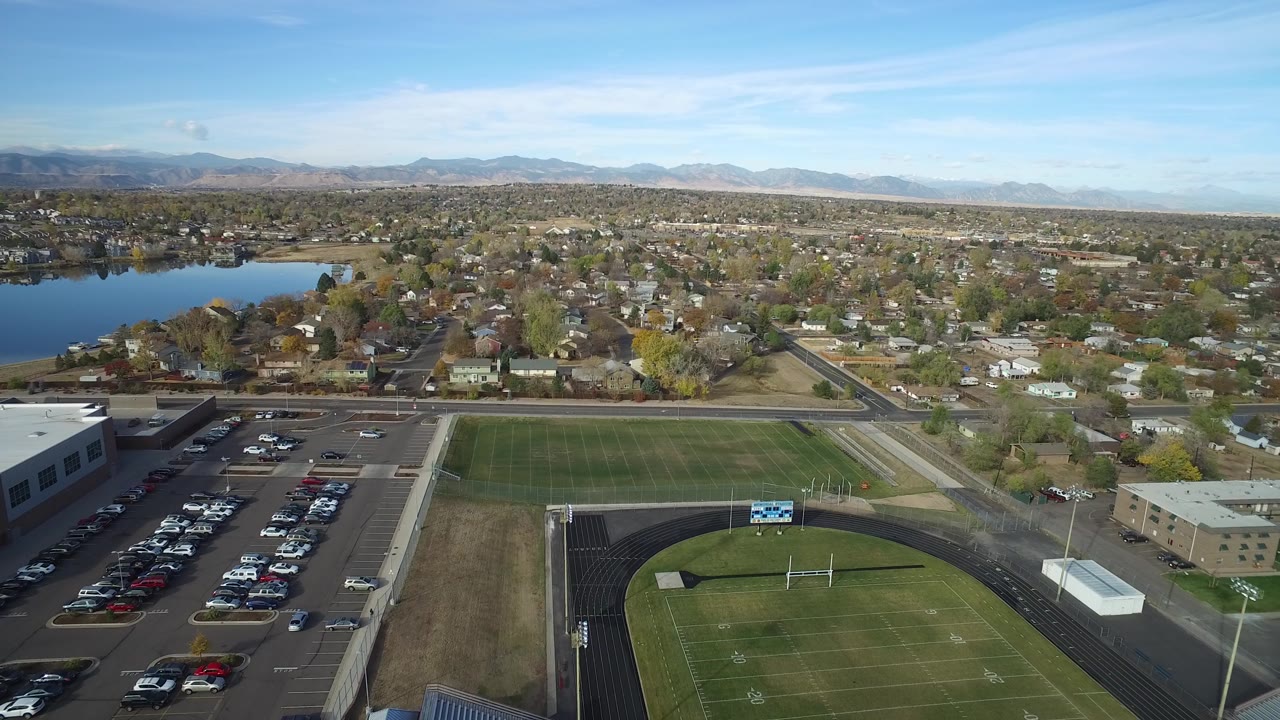 Aerial frame in Hidden Lake, Colorado with the football field centered lower in the view, a parking lot to the left, a tan low building to the right, homes beyond, and a lake at upper left.