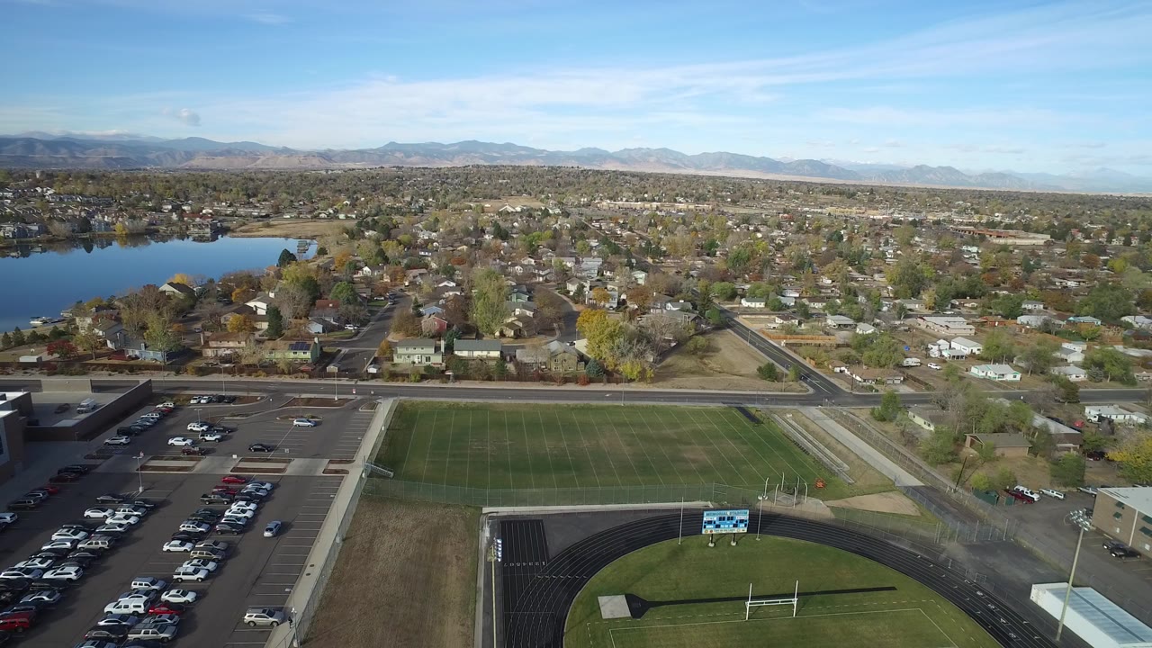 Wide aerial frame over Hidden Lake, Colorado with a running track and sports field in front, Glaze Lake on the left, suburban homes beyond, distant mountains, and no visible people.