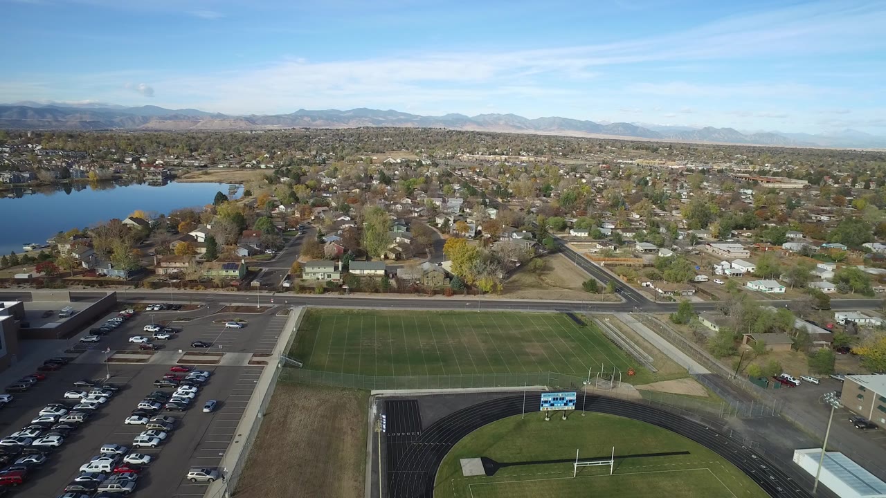 Aerial view in Hidden Lake, Colorado looking past a school-like field and parking lot toward Glaze Lake, residential streets, leafed trees, mountains, and no visible people.