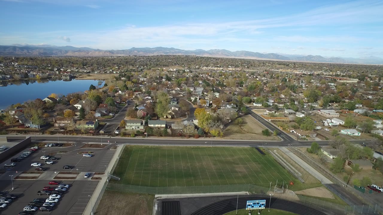 Aerial view in Hidden Lake, Colorado with a green field and parking lot at the bottom, homes and roads beyond, a lake on the left, distant mountains, and no visible people.