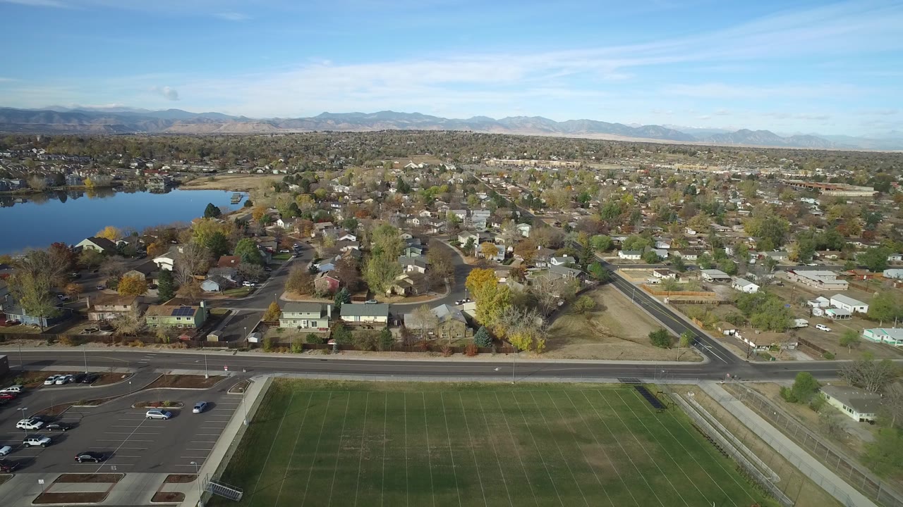 At 0:01, the Hidden Lake, Colorado view keeps the grass field and parking lot low in frame, with the lake left of center, rows of homes across the middle distance, mountains on the horizon, and no visible people.