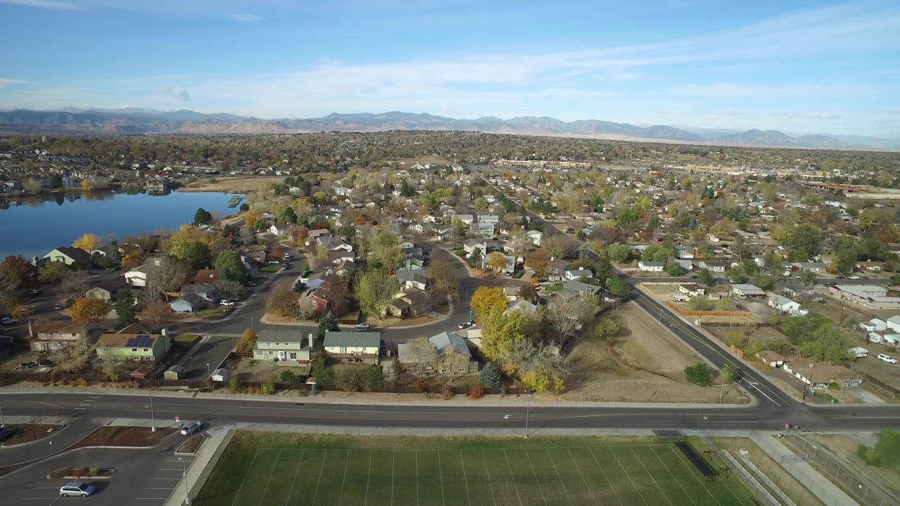 Opening aerial frame over Hidden Lake, Colorado, with a blue lake at left, neighborhood houses across the center, a road in the foreground, distant mountains, and no visible people.