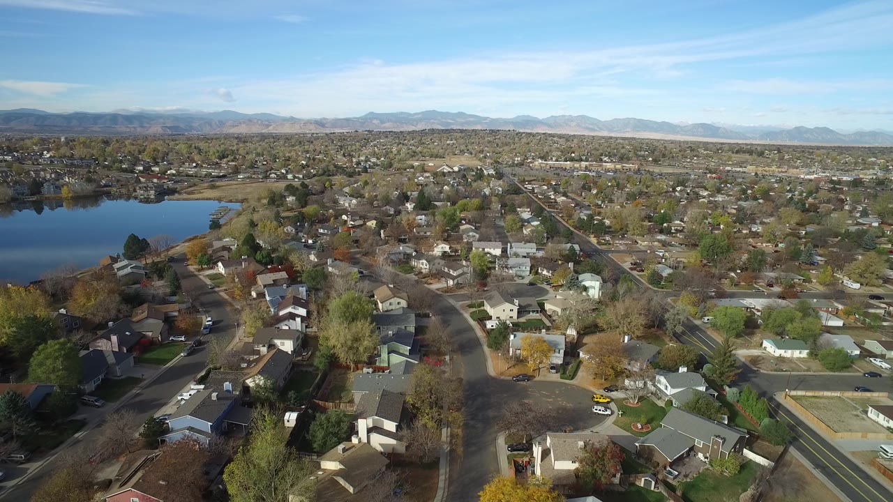 Aerial view over houses, streets, trees, lake water, and distant mountains.