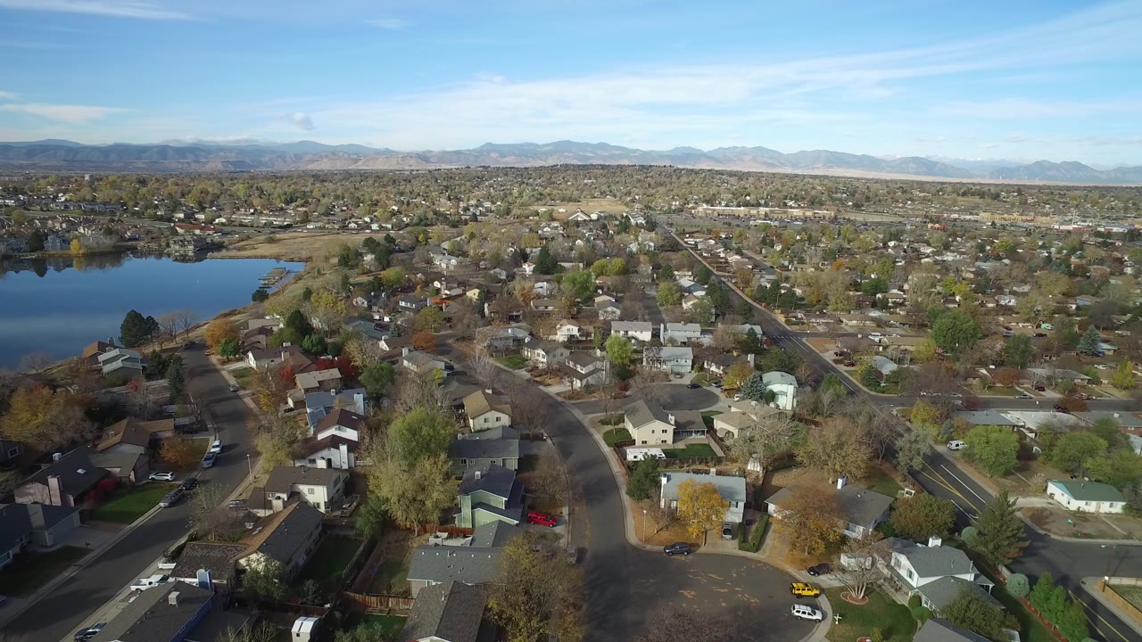High aerial view over Hidden Lake, Colorado homes and curved streets with the lake on the left, distant mountains on the horizon, and no visible people.