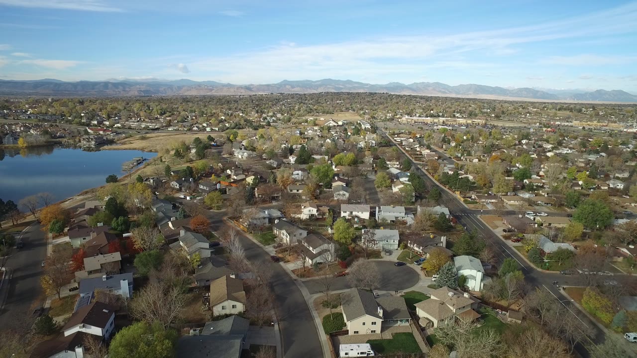 Opening aerial frame over Hidden Lake, Colorado, with suburban houses in the foreground, lake water on the left, distant mountains on the horizon, and no visible people.