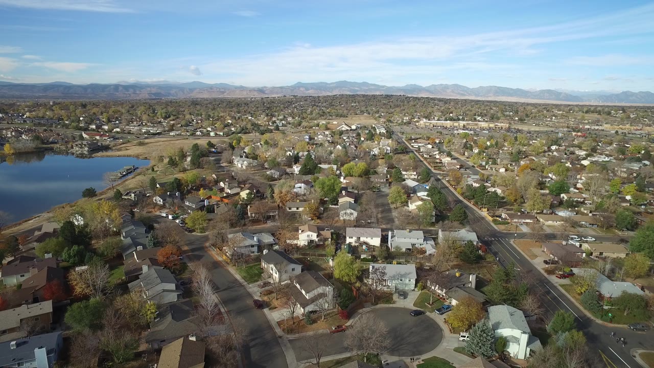 Second aerial frame in Hidden Lake, Colorado, showing more of the straight road through the neighborhood, the lake along the left edge, low homes, autumn trees, mountains, and no visible people.