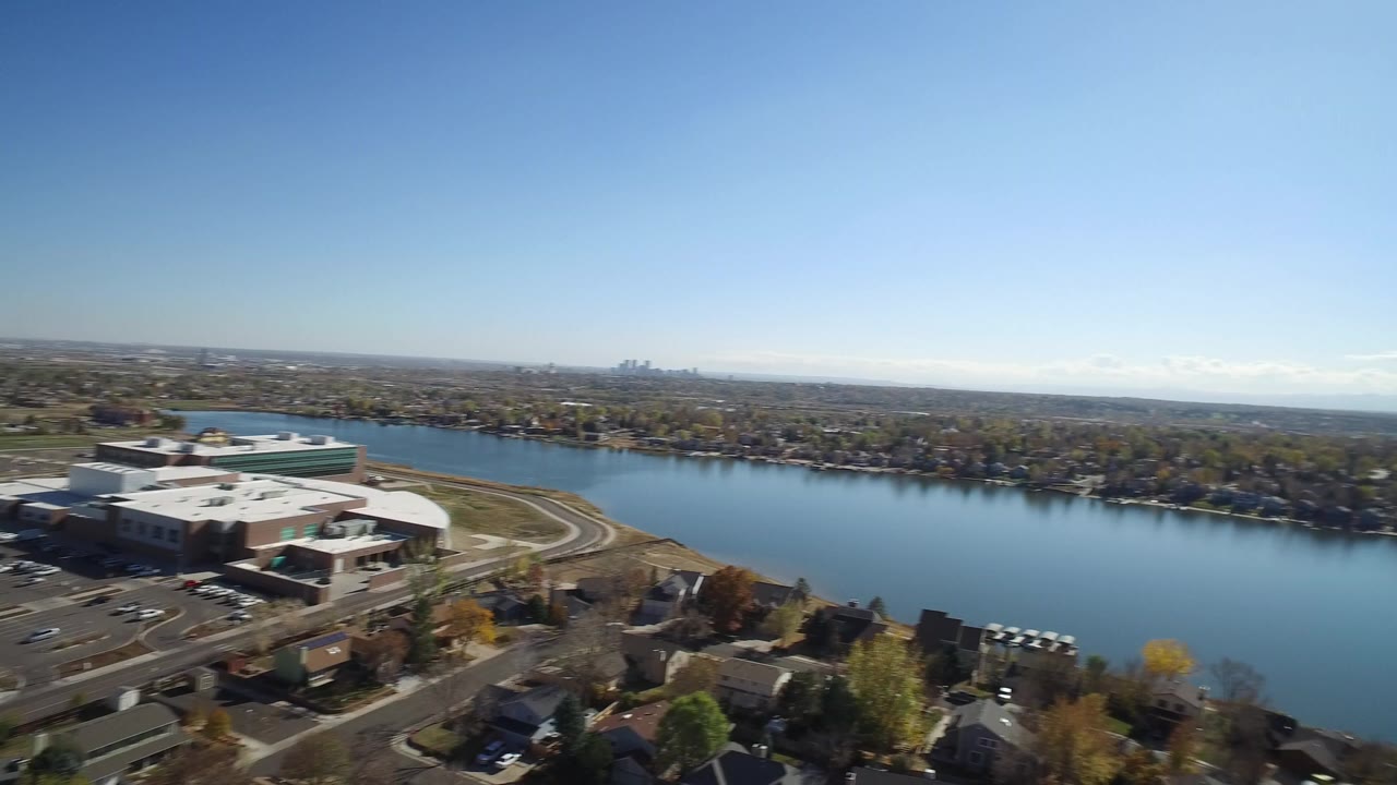 Opening aerial frame in Hidden Lake, Colorado showing a blue lake beside a large low building, nearby roads, residential roofs, distant city skyline, and no visible people.